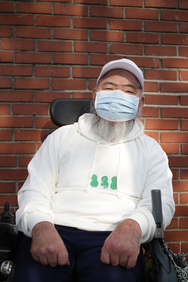 녹색 눈사람(흰색 후드) 을 입고 휠체어에 앉아 양손을 모은 포즈를 취한 노들장애인야학 교장선생님