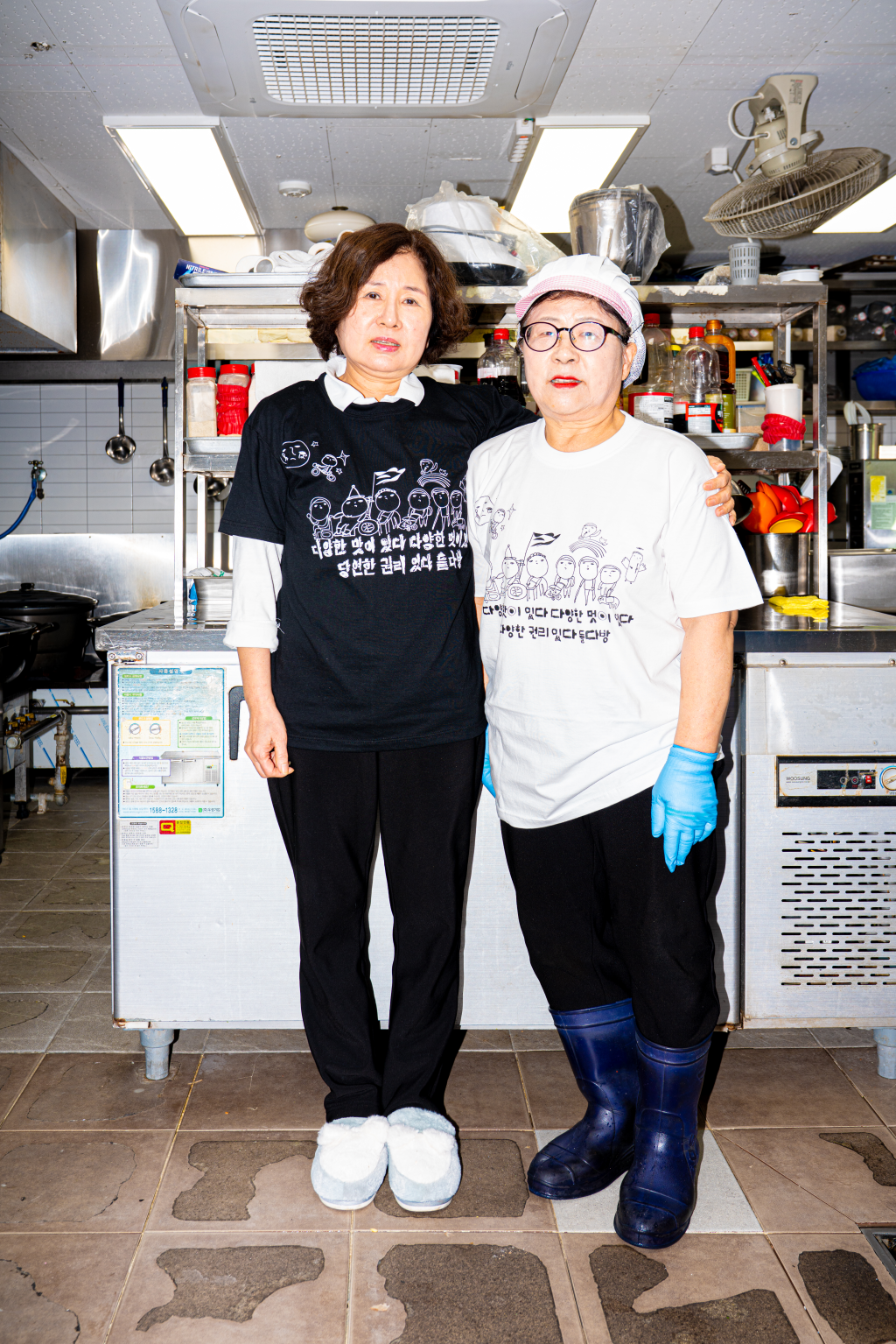 들다방 주방 내 조리대를 등지고 환하게 웃으며 이편을 보는 두 사람. 왼편 사람은 검정색 들다방 티셔츠를 입고 한 손으로 다른 한 사람의 어깨를 감싸 안았다. 오른편 사람은 조리 모자, 파란 위생장갑을 낀 채 흰색 들다방 티셔츠를 입었다. 세로로 긴 전신 사진.
