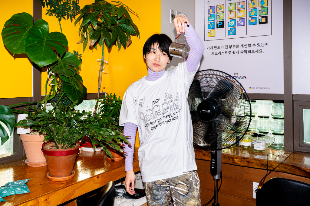 들다방 카페 벽면 화분 앞에서 들다방 흰색 티셔츠를 입고 포즈를 취한 모델 상반신 사진. 한손에 스텐 컵의 손잡이 대신 키링(고리) 부분을 들어 보이고 있다.