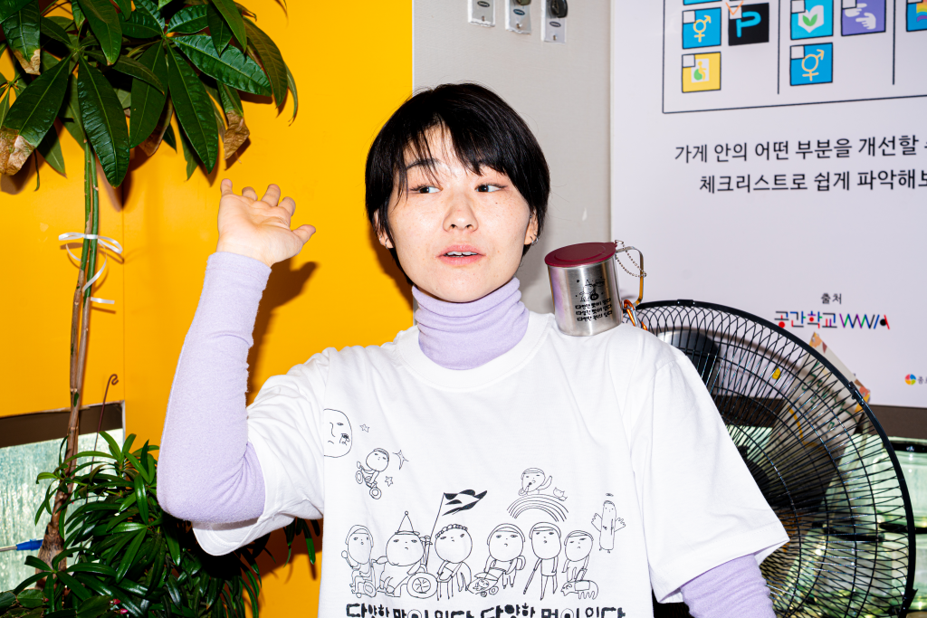 들다방의 노란색, 분홍색 벽면을 등지고 들다방 티셔츠 흰색을 입은 모델 상반신 샷. 한쪽 어깨에 들다방 스텐 컵이 놓여 있다