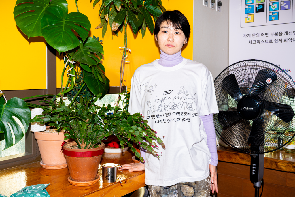 들다방의 노란색, 분홍색 벽면, 무성한 실내 초록 화분 잎사귀들 옆에서 들다방 티셔츠 흰색을 입은 모델 상반신 샷. 티셔츠 전면이 모두 드러나 있다.