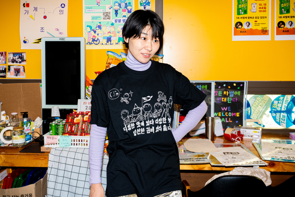 들다방 노란 벽면을 등지고 들다방 검정 티셔츠를 입은 모델 상반신 샷.