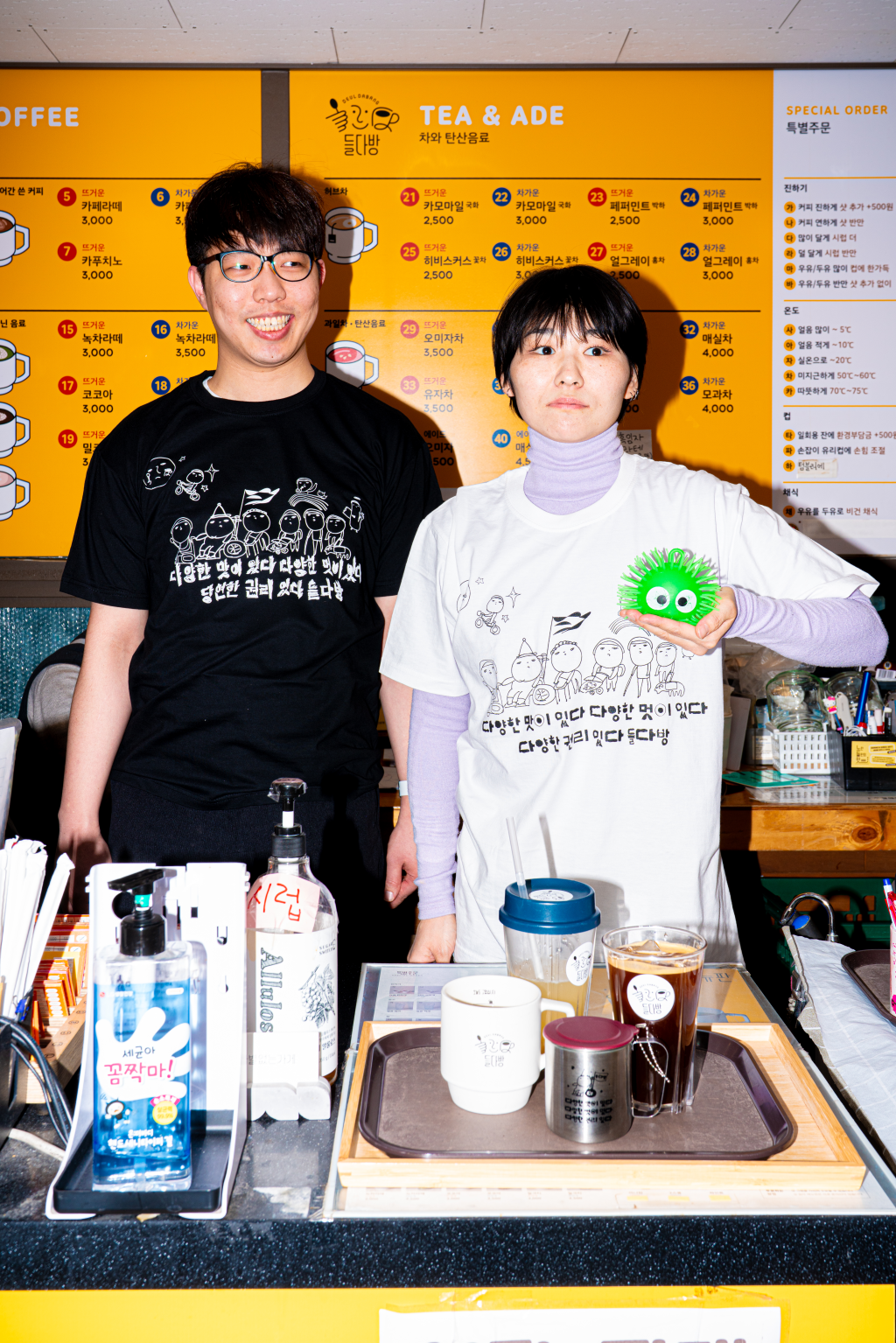 카페 들다방 벽면 메뉴판을 등지고 접수대 앞에서 들다방 티셔츠를 입은 두 사람. 왼편 사람은 검정색, 오른편 사람은 흰색 티셔츠 차림, 그들 앞 테아블의 쟁반 위에 들다방 스텐 컵과 함께 아이스, 핫 음료들이 준비돼 나와 있다.