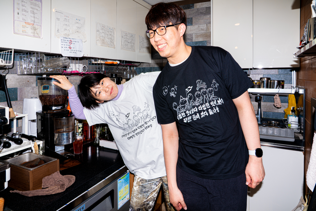 들다방 카페 조리 구역 내 에스프레소 머신 앞에서 포즈를 취한 두 사람. 조금 앞에 선 오른 사람은 들다방 검정 티셔츠를 입고 사선 앞을 바라보며 환히 웃고 있다. 조금 뒤켠에 선 왼쪽 사람은 흰색 들다방 티셔츠를 입고 원두 글라인더에 한손을 한손을 받치며 몸을 옆으로 기울인 채 장난스레 이편을 바라보며 웃는다.