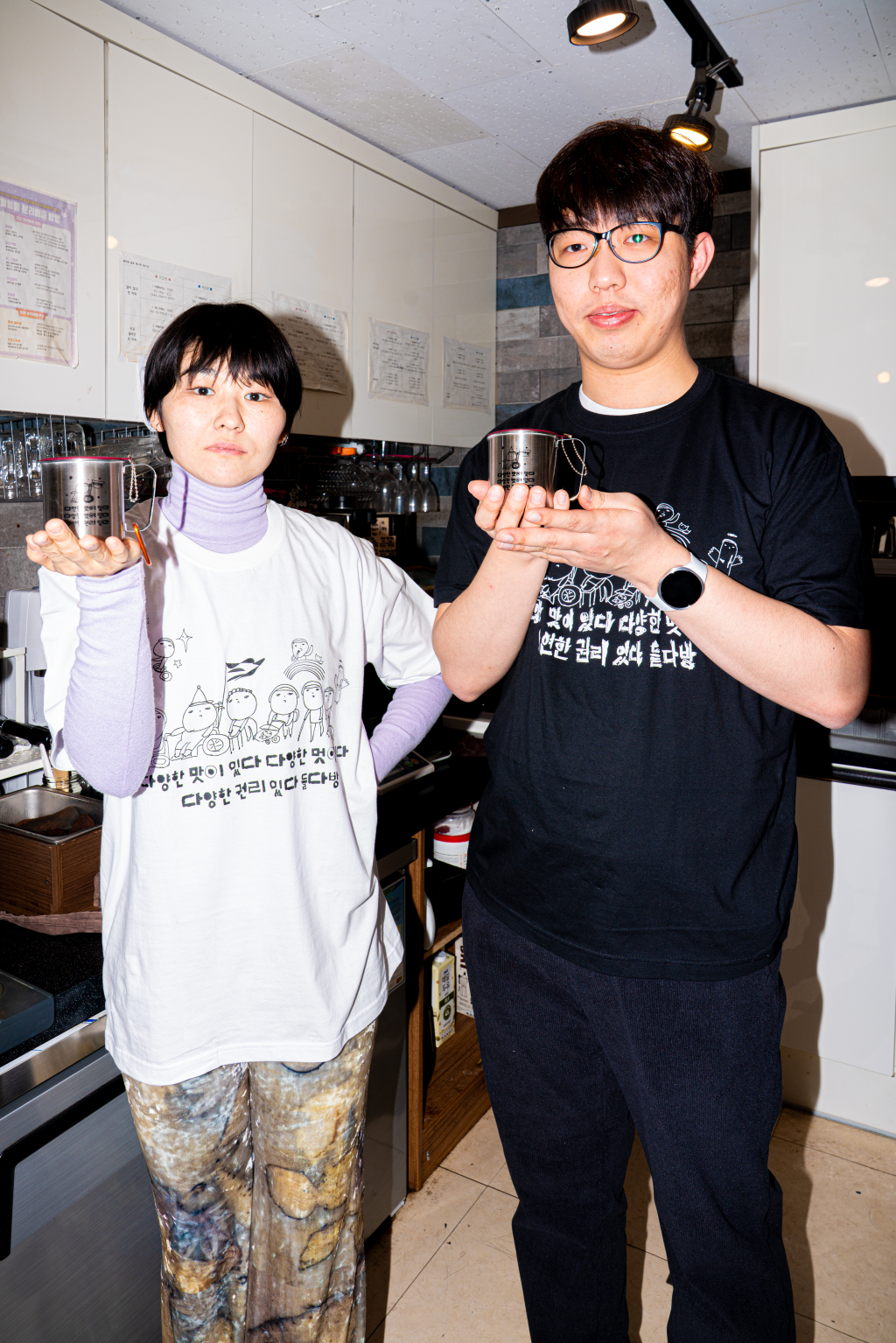 들다방 카페 조리 구역 내 에스프레소 머신 앞에서 포즈를 취한 두 사람 전신 샷. 왼쪽은 흰색 들다방 티셔츠를 입고 한손으로 들다방 스텐 컵을 들어 보이는 사람, 오른쪽은 들다방 검정 티셔츠를 입고 다소곳이 웃으며 양손으로 들다방 스텐 컵을 들어 보이는 사람.