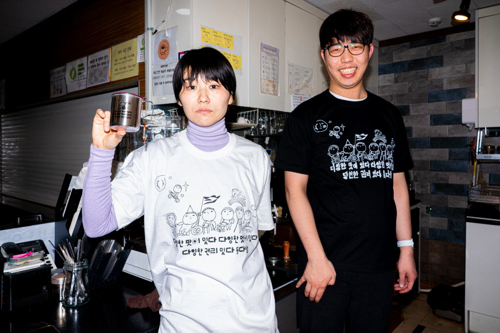 들다방 카페 조리 구역 내 에스프레소 머신 앞에서 포즈를 취한 두 사람. 왼쪽은 흰색 들다방 티셔츠를 입고 들다방 스텐 컵을 한손에 들어 보이고 있다. 오른쪽은 들다방 검정 티셔츠를 입고 이편을 보며 환히 웃고 있다.