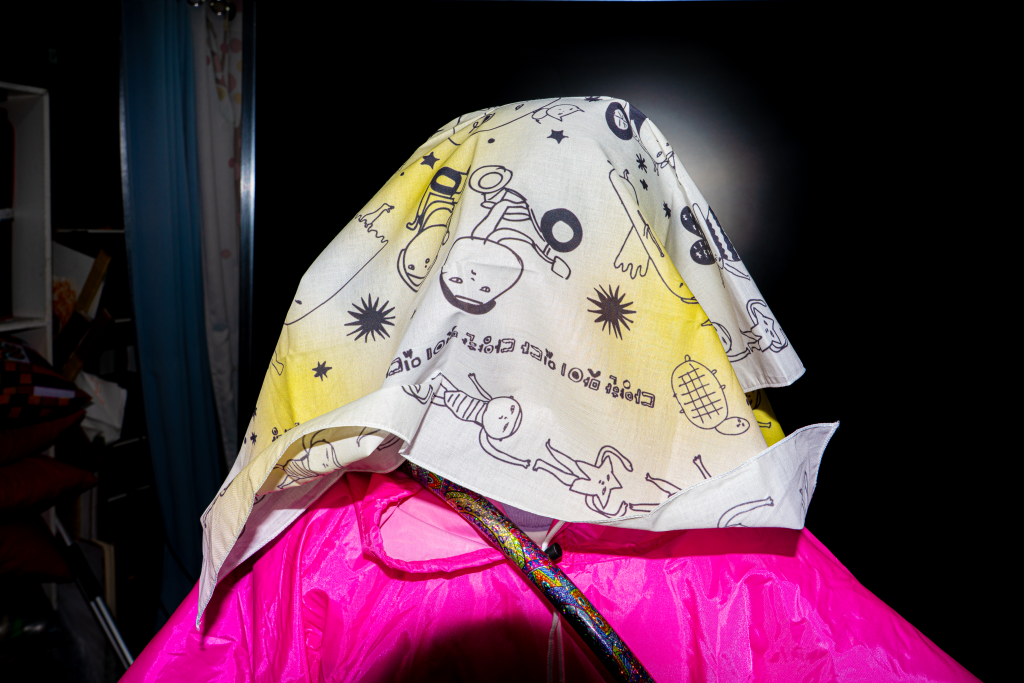 어두운 배경에 들다방 손수건을 머리에 얹어 얼굴을 모두 덮은 사람 숄더 숏. 들다방 핑크 판초 우비를 입고 있다.