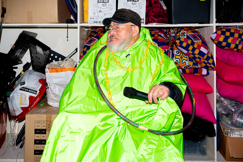 들다방 초록 판초 우비를 입은 채 한손에는 마이크를, 목에는 노란 사슬을 휘감은 사람 상반신 샷. 사선으로 멀리 앞을 바라본다.