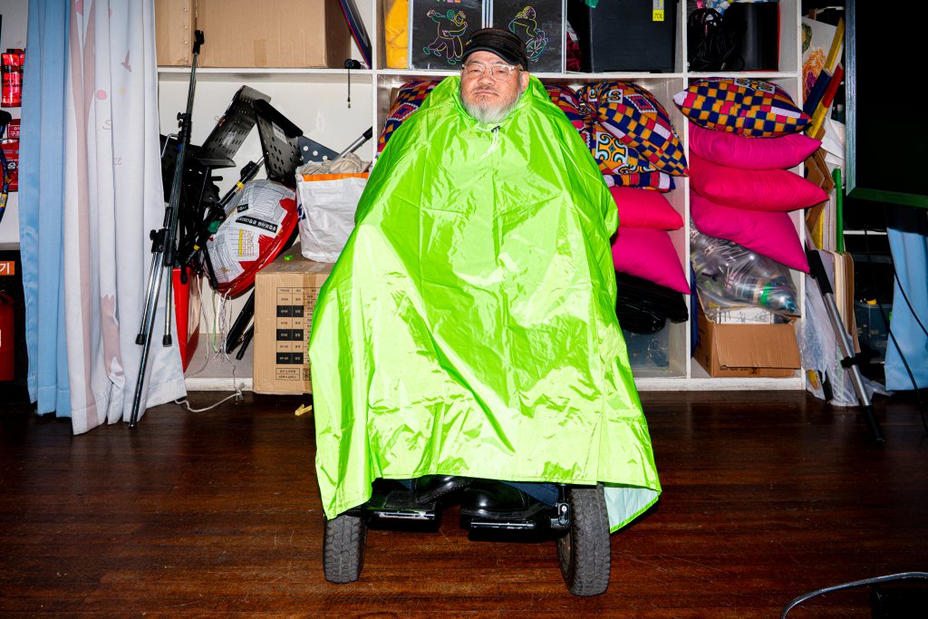 들다방 초록 판초 우비를 입은 채 전동휠체어에 앉아 있는 사람 전신 정면 샷. 머리 아래로 온몸이 우의 천으로 덮여 있으며 하단 신발과 지면에 닿은 두 바퀴만 드러나 있다.