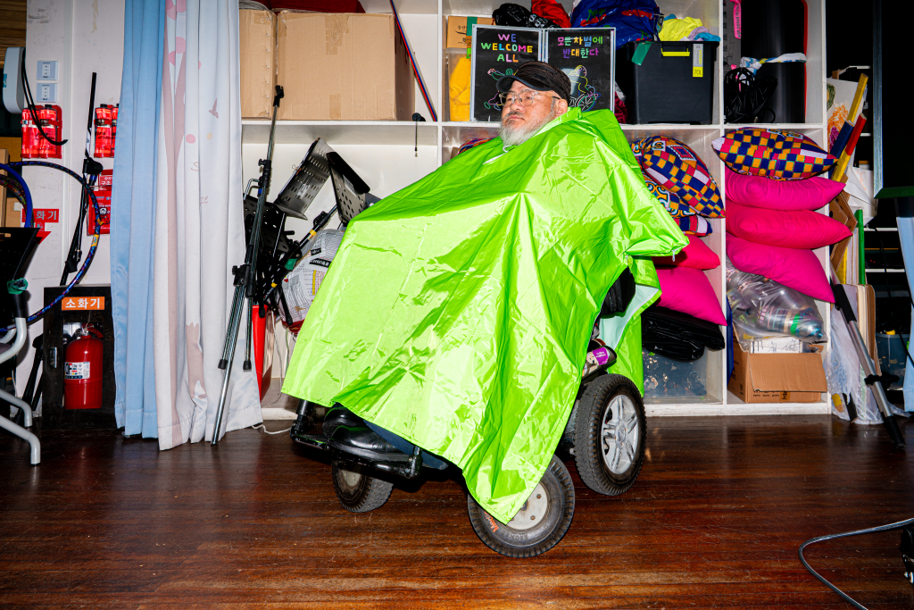 들다방 초록 판초 우비를 입은 채 전동휠체어에 앉아 있는 사람 전신 사선 샷. 판초 우비 측면 지퍼를 열어 넉넉하게 걸치니 머리 아래로 온몸이 우의 천으로 덮여 있다.