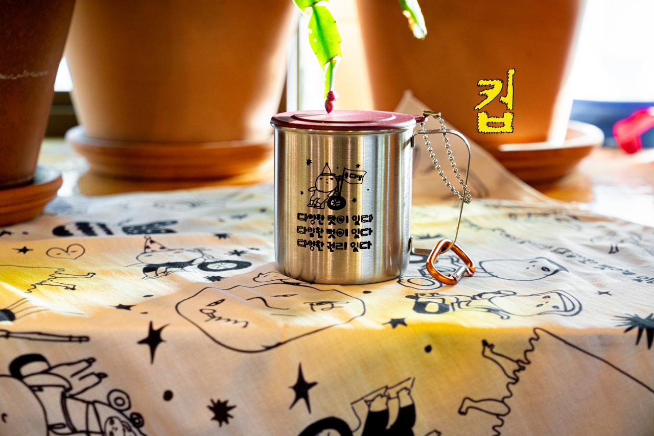 따뜻한 햇살을 받는 스테인리스 컵 클로즈업. 컵에는 그림과 글이 다음과 같이 프린트되어 있다. 들다방이라 쓰인 깃발을 휠체어에 달고 파티 모자를 쓴 채 나아가는 사람 그림. 하단에는 동글동글 가리런한 손글씨체로 ‘다양한 맛이 있다, 다양한 멋이 있다, 다양한 권리 있다 들다방’. 컵의 바닥에는 들다방 노랑 손수건이 깔려 있다.