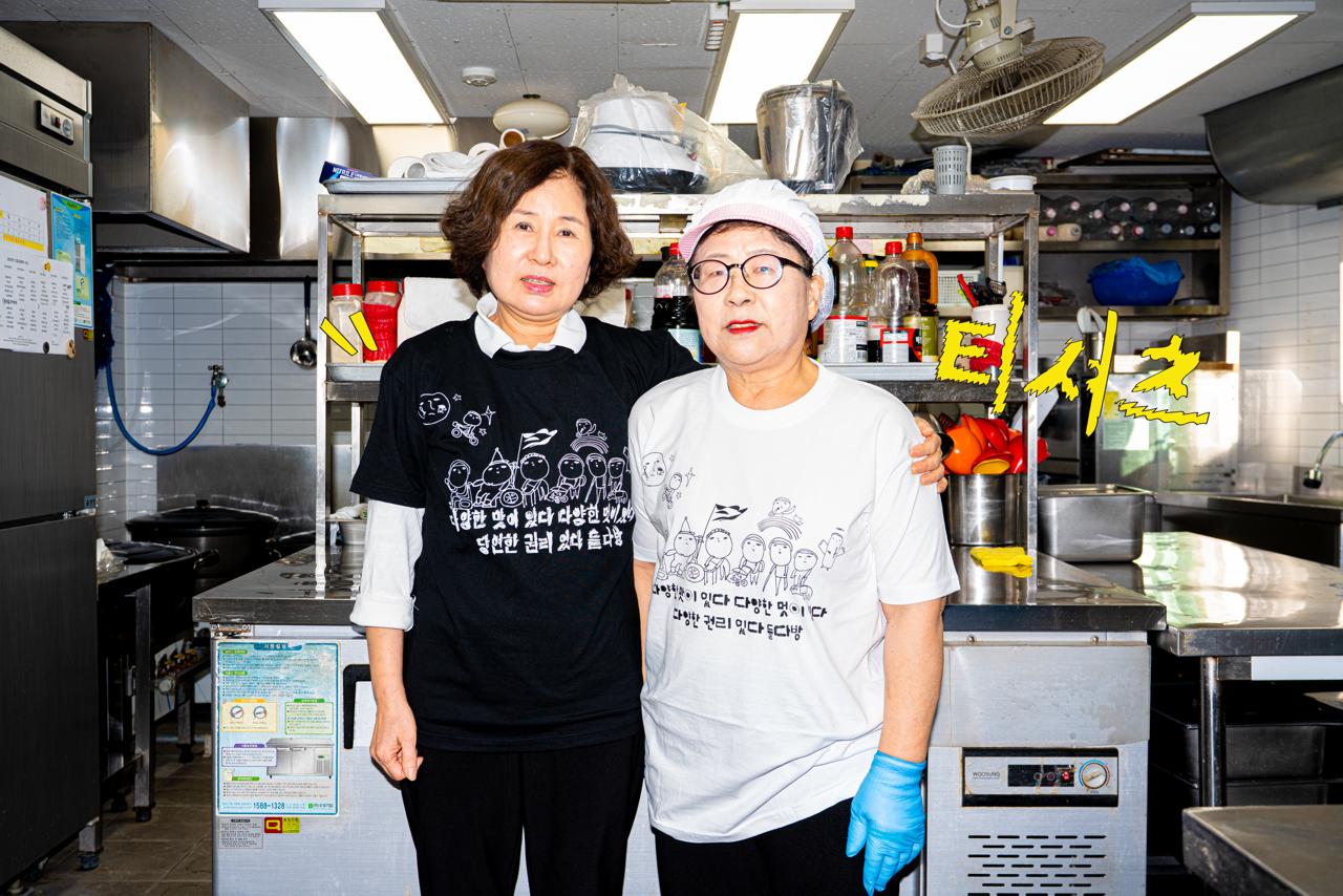 들다방 주방 내 조리대를 등지고 환하게 웃으며 이편을 보는 두 사람. 왼편 사람은 검정색 들다방 티셔츠를 입고 한 손으로 다른 한 사람의 어깨를 감싸 안았다. 오른편 사람은 조리 모자, 파란 위생장갑을 낀 채 흰색 들다방 티셔츠를 입었다.