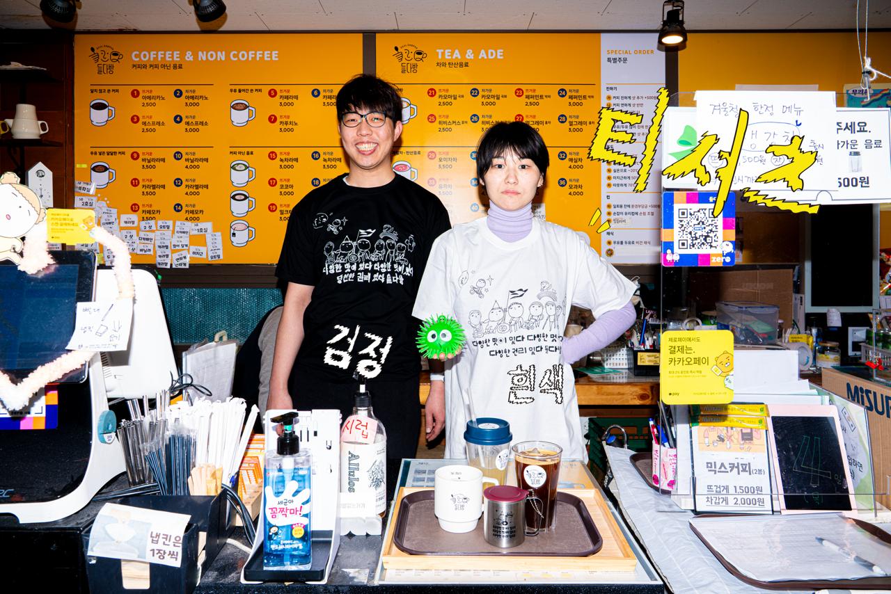 카페 들다방 벽면 메뉴판을 등지고 접수대 앞에서 들다방 티셔츠를 입은 두 사람. 왼편 사람은 검정색, 오른편 사람은 흰색 티셔츠 차림이다.