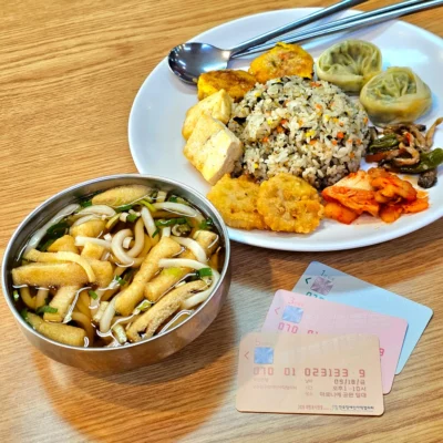 유부가 들어간 우동 대각선 위쪽 옆에 동그란 흰 접시에 동그랗게 담긴 볶음밥을 둘러싸고, 시계방향으로 찐만두, 버섯꽈리고추볶음, 김치, 연근튀김, 구운 두부, 동그랑땡이 담겨있다. 그리고 우동 우측 옆에는 전장야협 티켓 3종이 나란히 놓여있다.