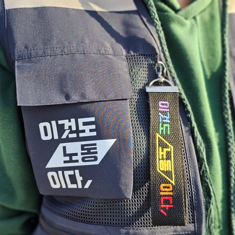 검은 조끼 주머니에 '이것도 노동이다' 문구가 인쇄되어 있고, 옆에는 무지개색 글씨로 이것도노동이다라고 적힌 키링이 달려 있다.