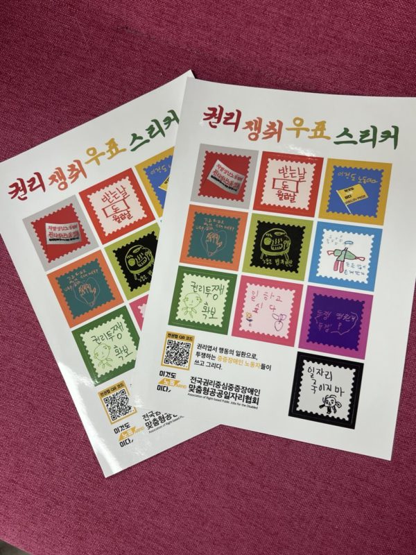 분홍색 천 위에 ‘권리 쟁취 우표 스티커’ 두 장이 놓여 있다. 중증장애인 노동자들이 직접 쓰고 그린 노동권·권리투쟁 문구를 우표 모양의 다채로운 스티커로 구성했으며, 전국권리중심중증장애인맞춤형공공일자리협회와 QR코드가 표시돼 있다.